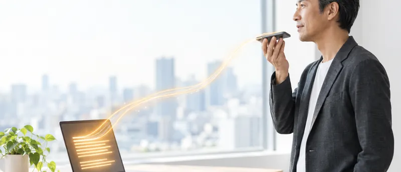 窓辺でスマートフォンに音声メモを吹き込むコンサルタント。声が琥珀色の光になってノートPCの画面に原稿として流れ込む